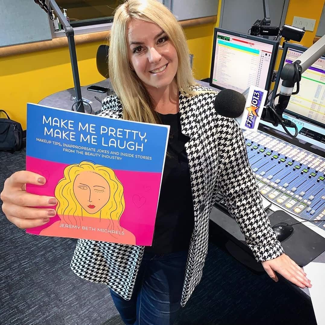 blonde woman in a radio studio holding up a copy of Make Me Pretty, Make Me Laugh