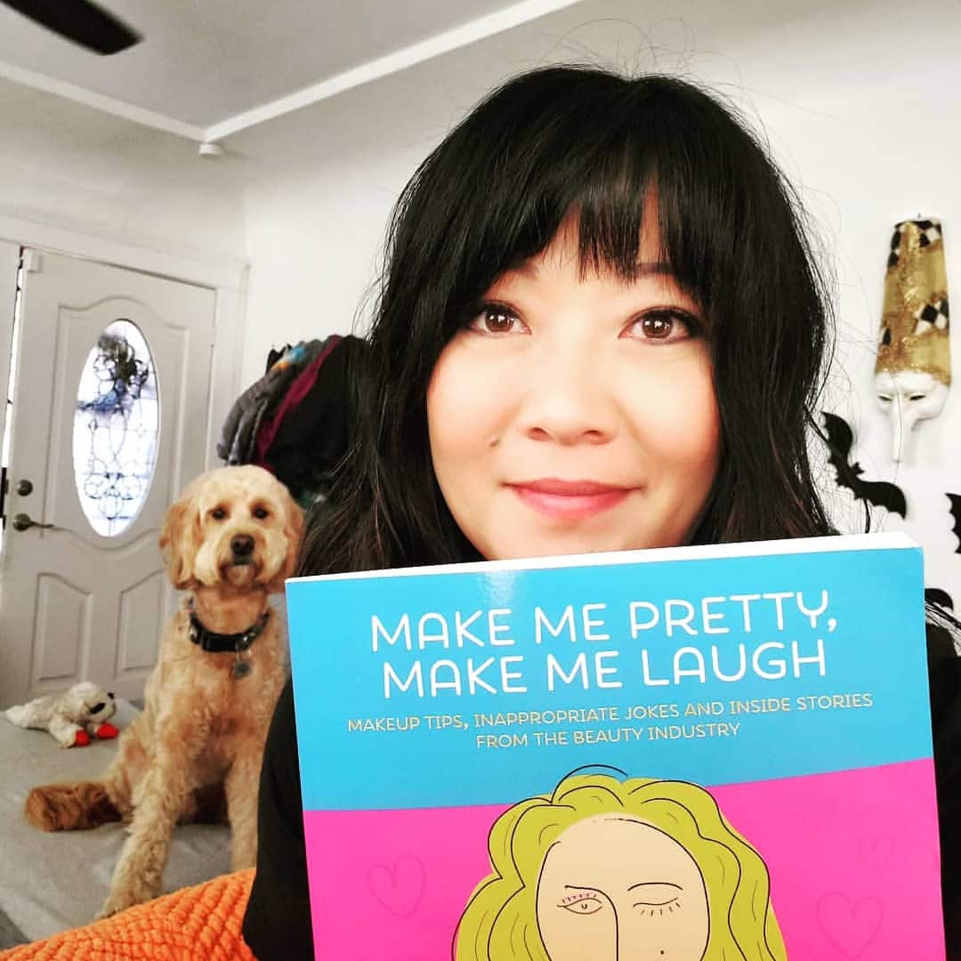 woman with dog in background holding up a copy of Make Me Pretty, Make Me Laugh
